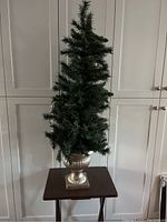 Full view of one artificial Christmas tree on a small wooden table, with urn base and warm string lights on tree branches.