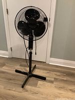 Full view of the black floor fan showing its stand and base on a wood floor.