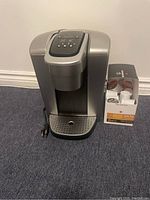 Silver Keurig coffee machine with power cord shown next to box of coffee pods.