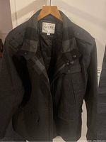 Front and interior view of men's Guess winter jacket hanging on a wooden hanger, showing gray exterior with flap pockets and black quilted lining inside.