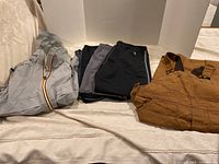 Overall view of all clothing items laid out: light gray rain suit, multiple pants in different colors and brands, and brown overalls.