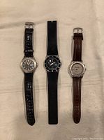 Three men's wristwatches laid flat on a fabric surface, showing full watches including straps and faces.