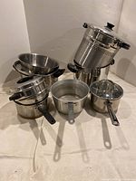 Photo showing assorted stainless steel cookware including two saucepans, pasta strainer, and steamer basket on a light background.