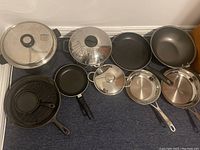 Full overhead view showing all pots and pans laid on floor, including cast iron pans, stainless steel pots with lids, non-stick pans, steamer insert, and brie baker dish.