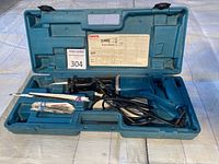 Makita reciprocating saw inside opened original blue plastic carrying case with blades in compartments and loose inside, showing tool and blades arrangement.