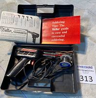 View of opened hard black plastic carrying case containing soldering gun, solder wire spool, power cord, and instructional booklets.