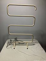 Front view of the off-white S-shaped freestanding electric towel warmer positioned on a white sheet against a gray wall.