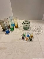 Full lot showing tall colored glasses, small colored shooters, clear shooters, and green glass jar on a white patterned tablecloth.