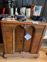 Front view of antique wood cabinet showing two central curved and carved doors with metal knobs, flanked by lighter wood panels, topped with various small household objects.