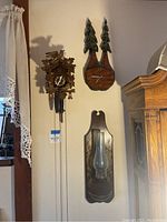 Photo showing three wall mounted items: a carved wooden cuckoo clock with pinecone weights, a carved wooden tree shaped clock, and a wooden candle holder with glass chimney with floral painted decoration.