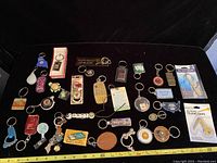 Photo showing a large assortment of souvenir key chains and tools laid out on a black fabric background, including various promotional and souvenir designs.