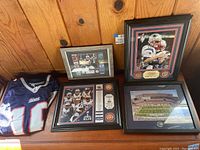 Photo showing the Patriots number 16 jersey laid out beside four framed pieces of Patriots memorabilia including Tom Brady, Rob Gronkowski, Gillette Stadium, and a 2015 championship collage.
