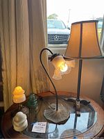 Three lamps on a small round table beside a window, showing the curved metal base lamp with ruffled glass shade lit, alongside beige lamp and antique glass insulators.