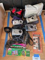Overview of cameras and accessories laid on floor, showing all items in the lot including various camera models, cases, film pack and LensPen.