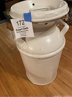 Side view of white painted metal milk jug with collar lid, side handles, and embossed text.