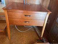 Front angle showing tapered legs, two drawers with metal handles, and teak wood grain on the table.
