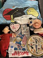 Photo showing a variety of vintage cartoon memorabilia items including Popeye plush, Betty Boop tin, Bugs Bunny phone book, Garfield welcome cloth, and others.