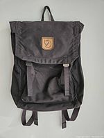 Front view of black Fjallraven waxed backpack hanging against a white wall, showing flap top with straps and leather logo patch.