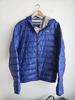Front view of blue Lacoste men’s down jacket hanging on a hanger showing full jacket with visible zipper and hood