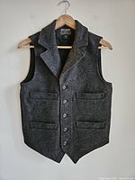 Front view of the dark gray Filson wool vest hanging on a wooden hanger against a white wall, showing button front and three pockets.