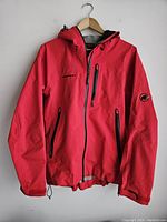 Full front view of the red Mammut jacket hanging on a wooden hanger, showing three zippered pockets and adjustable hood and cuffs.