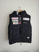 Front view of black Descente men's puffer vest with zipper, hood, and multiple sponsor patches including Raiffeisen, Helvetia, BKW, Swisscom, and Descente logo.