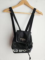 Front view of the vintage POLO SPORT black backpack hanging on wooden hanger, showcasing front flap, drawstring and strap details.