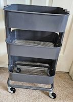 Front angle showing full 3-tier black rolling cart with mesh tray bottoms and caster wheels on carpeted floor.
