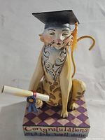 Front view of the Jim Shore cat figurine wearing a graduation cap and holding a diploma on a purple diamond-patterned base