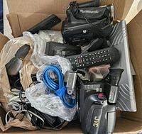 Box containing two vintage camcorders, remote control, cables, Apple charger, and other electronic accessories.