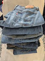 Stack of seven pairs of Levi's jeans in different shades of blue denim, showing back pockets and leather patches on some.