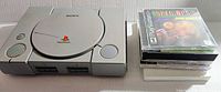 Gray Sony Playstation 1 console with six games stacked beside it, showing console buttons and ports.