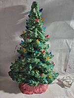 Front view of a green ceramic Christmas tree with multicolored glass bulbs on a red base with attached power cord.
