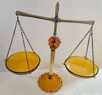 Front view of the amber glass Scale of Justice balanced with two round hanging plates on chains attached to a metal frame with an amber glass base and ornamental orb.
