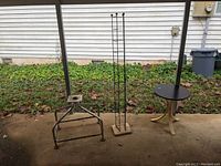 Three pieces of outdoor furniture including two table bases and one tall narrow metal stand arranged on concrete patio outside with grass and siding in background.
