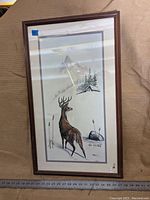 Framed print showing full artwork of buck standing in winter mountain scene, signed at bottom right.