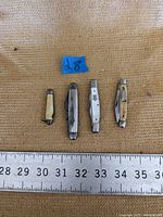 Four collectible folding pocket knives laid out with measuring ruler showing sizes ranging from about 2.5 to 3.5 inches closed on a burlap surface.