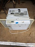 Top view of GE window air conditioner laid flat on a wooden surface showing control panel, remote control placed on top, power cord coiled, and window mounting panels attached to sides.