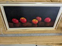 Framed painting showing five red apples on black background with a tape measure at the bottom indicating width.