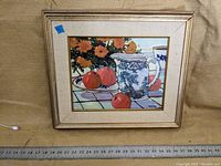 Framed still life painting showing orange flowers, a blue and white pitcher, and three red fruits on a tiled surface, with the artist's signature 'Mota' visible.