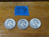 Three vintage silver Washington quarters arranged side by side on a wooden surface with a blue tape showing '89' above one coin.