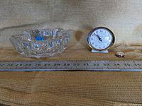 Clear glass bowl, clock-style watch, and small ring arranged next to a ruler for scale, on textured fabric surface.