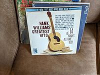 Two Hank Williams vinyl records with detailed album covers; 'Greatest Hits' featuring a guitar and hat illustration, 'Very Best of Hank Williams' with a drawn portrait.