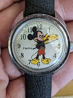 Close-up of the vintage Mickey Mouse electric watch by Timex, showing Mickey Mouse on the face with his hands as the hour and minute indicators. The watch strap is black leather with signs of wear.