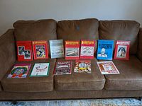 Photo showing 11 vintage circus magazines arranged on a brown couch with various colorful and black and white covers, some with illustrations of performers, animals, and posters.
