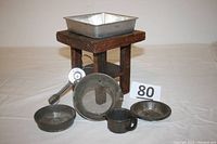 Full lot arranged on surface showing all items: rotary eggbeater, small wooden table, sieve, pie plate, round and square trays, cup