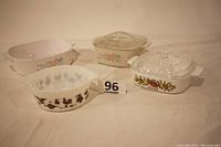 Set of four vintage casseroles and bowls arranged on white surface. Left to right: Pyrex Early American pattern bowl with brown farm animal silhouettes, CorningWare Lechalote rectangular casserole with fruit and vegetable decoration and clear lid, Corelle Symphony casserole in white without lid, Corelle Symphony casserole cream color with clear lid.