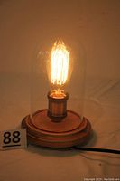 Lit Edison bulb inside glass dome on wooden base, showing warm filament glow and cord with switch.