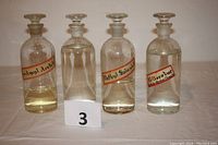 Photo showing four vintage glass apothecary bottles labeled Isoamyl Acetate, Methyl Salicylate, Cellosolve, and Phenolphthalein, with glass stoppers and liquid contents.