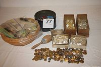 Wide shot showing multiple boxes of buttons, a wicker basket containing bags of buttons, a small metal pail, and a wooden tool on a white surface.